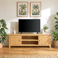 A light natural wood TV stand with cabinet doors and open shelving, styled in a living room setting with a black flat-screen TV on top, framed botanical prints on the wall, and potted plants on both sides.