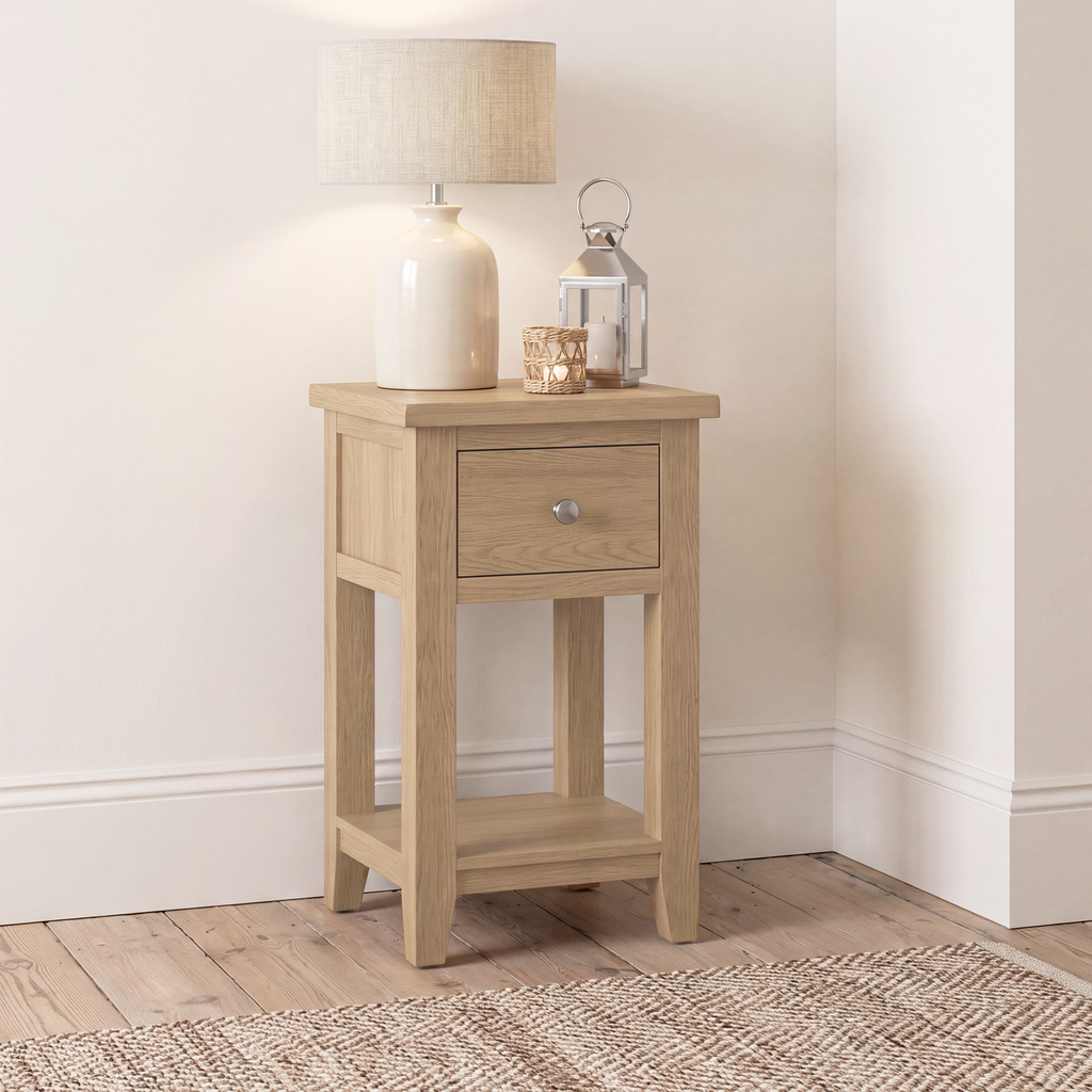 A light oak wood end table with a single drawer and open lower shelf, styled with a ceramic table lamp and lantern in a bright room corner.