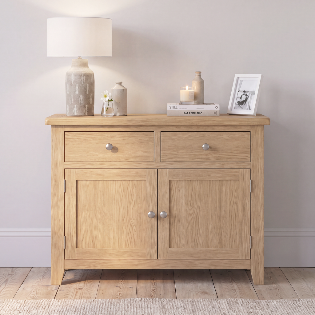 A light oak wood sideboard cabinet with two drawers on top and two doors below, styled with a table lamp, small vases, candles, and a framed print against a neutral wall.