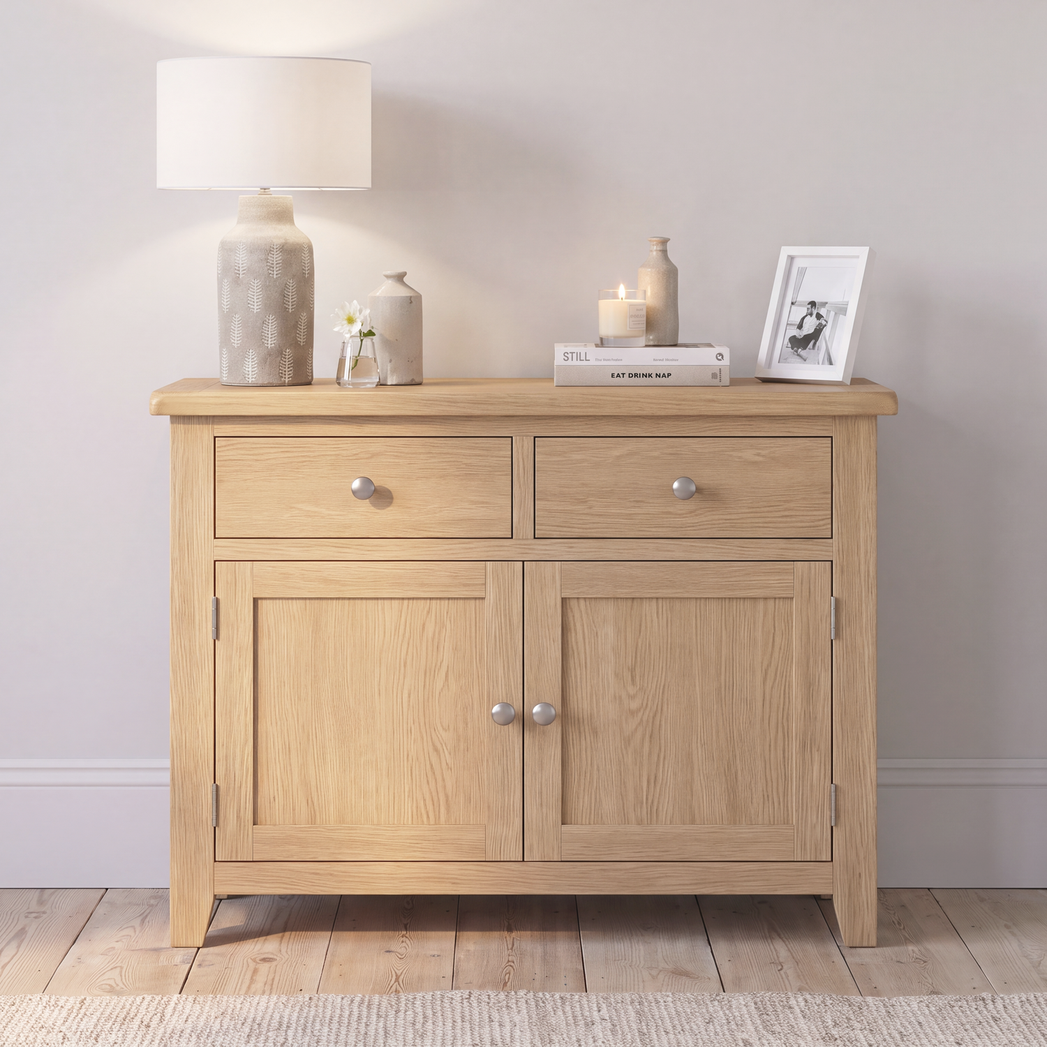 A light oak wood sideboard cabinet with two drawers on top and two doors below, styled with a table lamp, small vases, candles, and a framed print against a neutral wall.