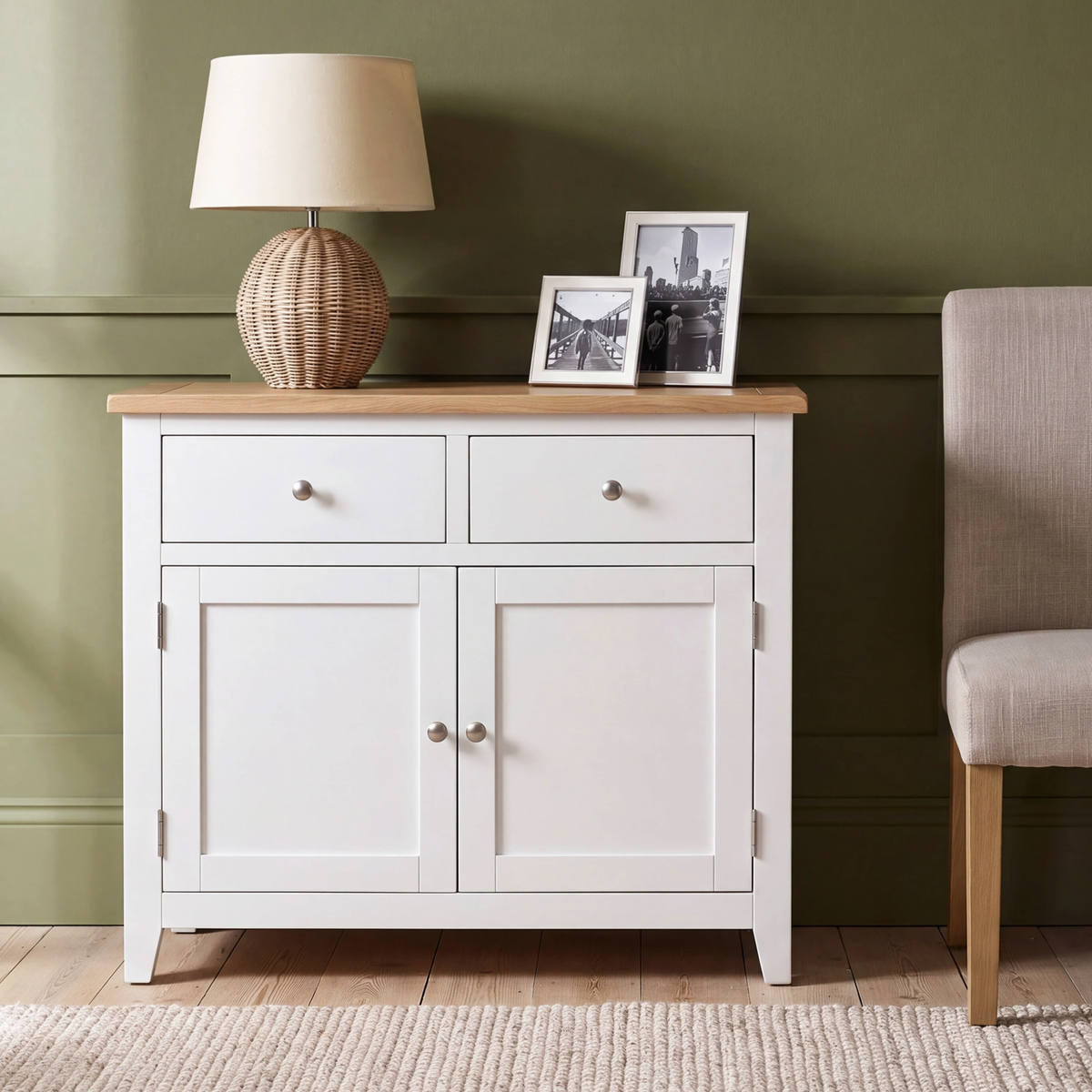 A white wooden sideboard cabinet with two drawers and two doors, styled in a room setting with a lamp, picture frames, a green wall, and a chair.
