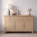 A light oak wood sideboard cabinet with three drawers on top and three paneled doors below, styled with a table lamp, framed photo, bowl, candle, and vase against a neutral wall.