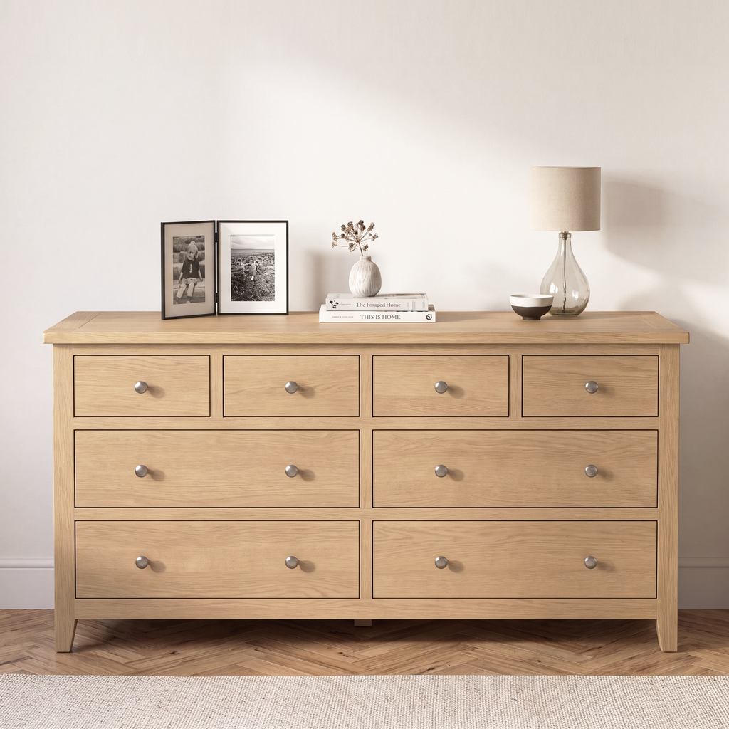 A light oak wood 8-drawer dresser with silver knobs, styled with picture frames, a vase, books, and a table lamp against a neutral beige wall.