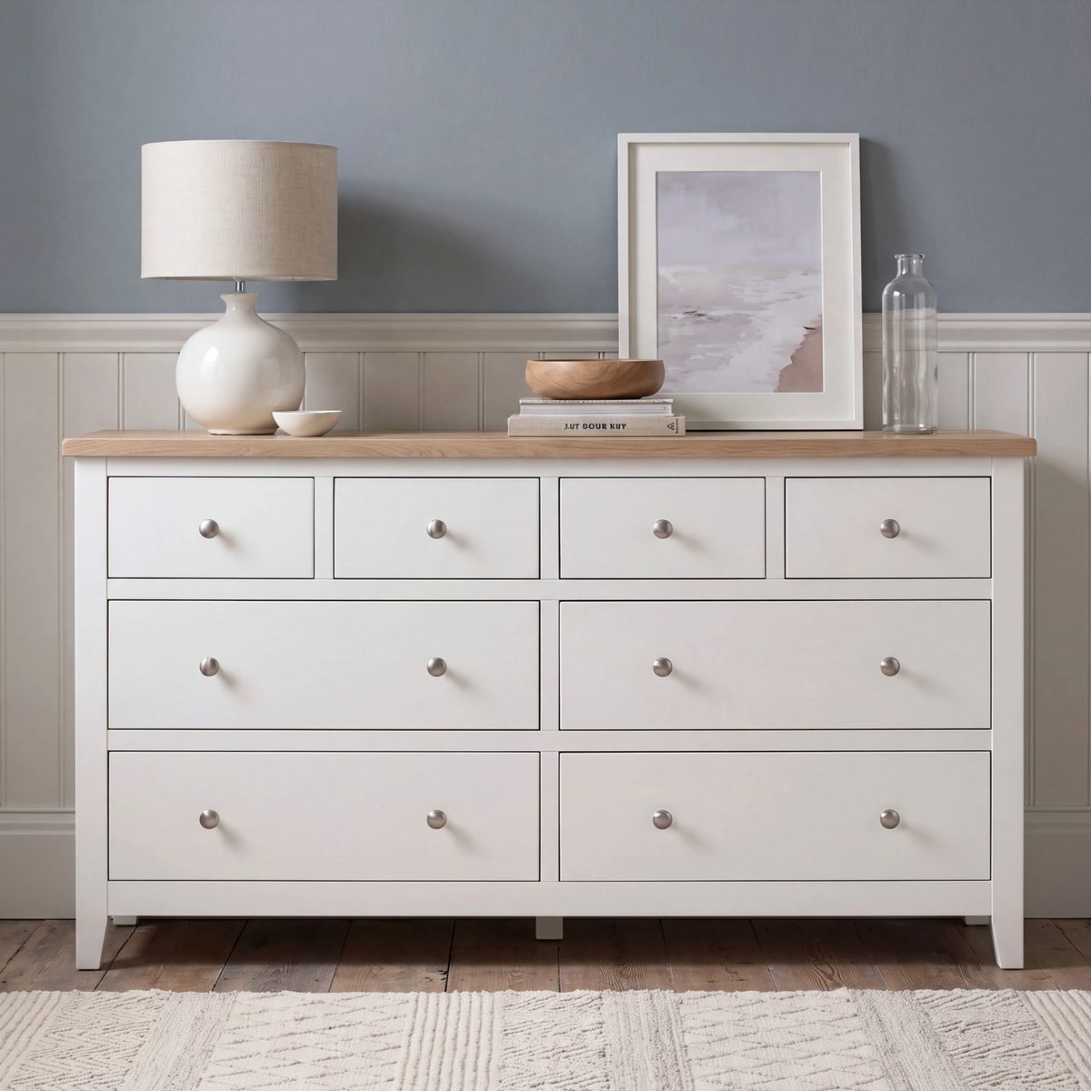 A white wooden dresser with eight drawers and small silver knobs, topped with a light natural wood surface. The dresser is styled in a bedroom setting with a white ceramic lamp, a framed print, books, a small bowl, and a clear glass bottle.