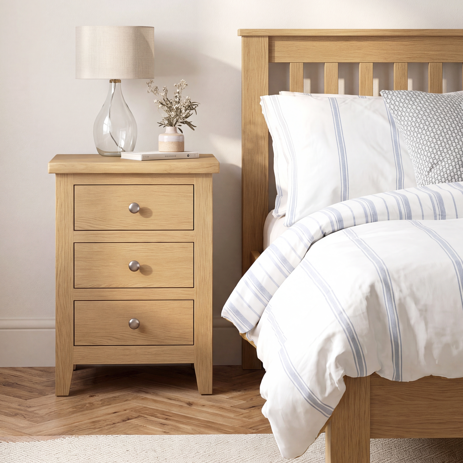 A light oak three-drawer nightstand beside a wooden bed frame with white and blue striped bedding, a table lamp, and a small vase in a bright bedroom setting.
