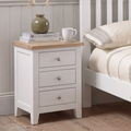 A white three-drawer nightstand with a light natural wood top, styled in a bedroom next to a white bed frame. The nightstand is decorated with a green ceramic vase, books, and a white mug.