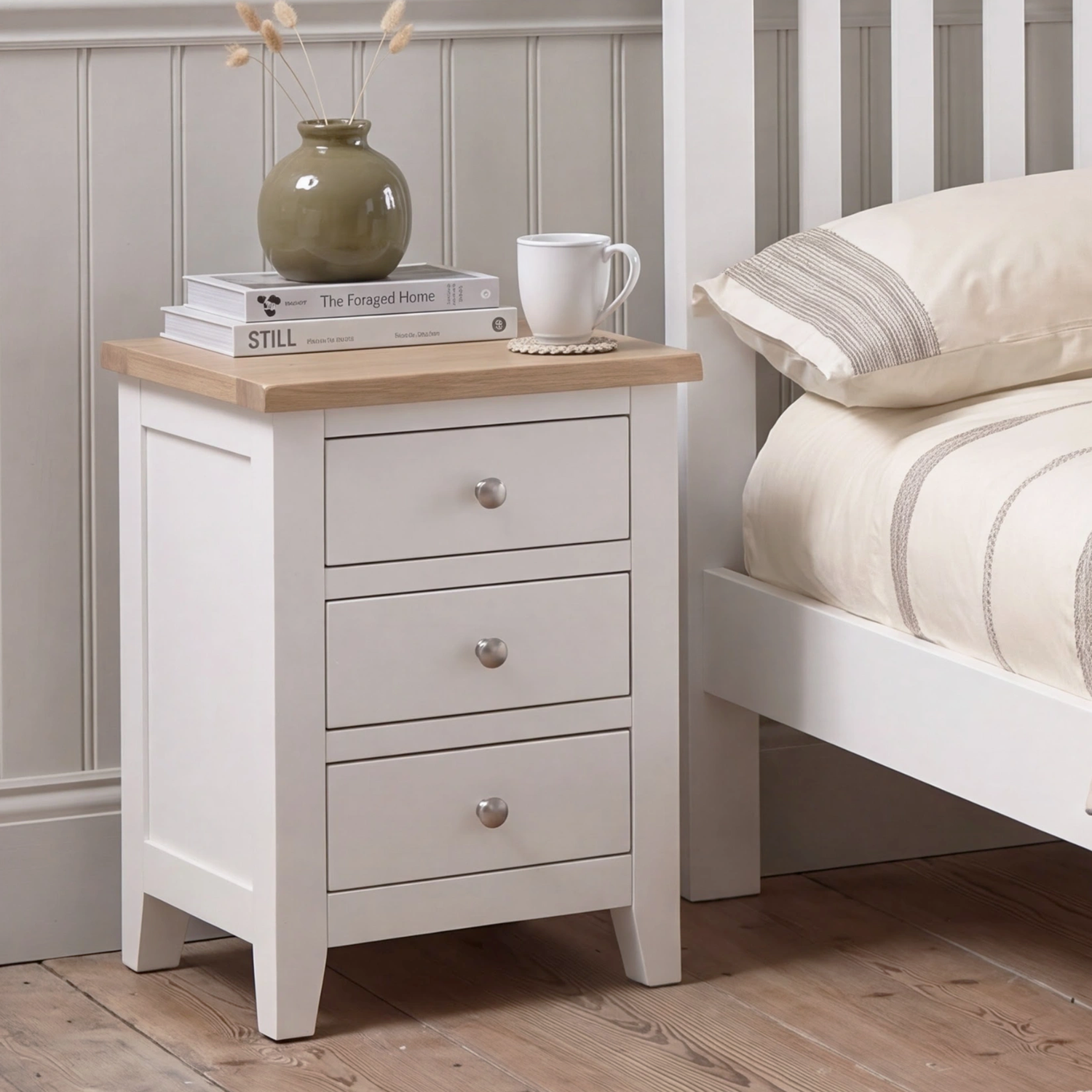 A white three-drawer nightstand with a light natural wood top, styled in a bedroom next to a white bed frame. The nightstand is decorated with a green ceramic vase, books, and a white mug.