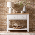 A white console table with a light wood top, two drawers, and an open lower shelf, styled with a table lamp, ceramic vases, and a woven basket in front of a rustic stone wall.