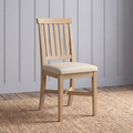 A light natural wood dining chair with a slatted backrest and a cream upholstered seat, shown standing on a woven jute rug against a gray paneled wall.