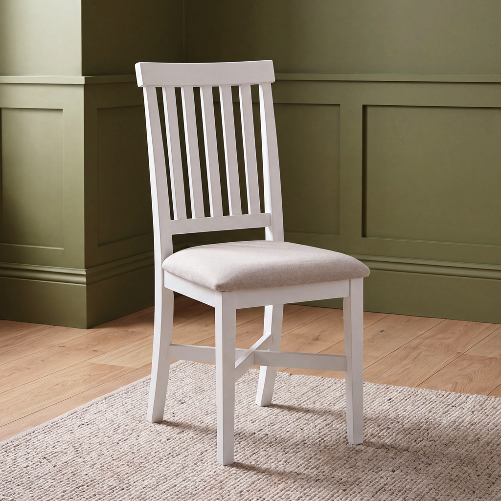 A white wooden side chair with a slatted backrest and a light beige upholstered seat, shown in a room with olive green paneled walls and a light wood floor.