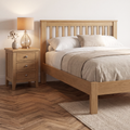A light oak wood bed frame with a slatted headboard, styled with white and beige bedding, next to a matching three-drawer nightstand with a glass table lamp, set on a herringbone wood floor.
