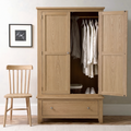 A light oak wood wardrobe with two doors open, showing hanging white and beige shirts, with a bottom drawer. A matching light oak wooden chair sits to the left against a neutral wall with a framed print.
