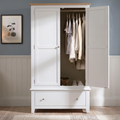 A white wooden three-door wardrobe with a light natural wood top and silver handles, shown open with clothes hanging inside and a single drawer at the bottom, set against a blue-gray wall with white wainscoting.