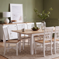 A white dining room furniture set with a rectangular light oak wood table and six matching white chairs with light wood seats, styled in a dining room with a green wall, white sideboard, and a vase of greenery.