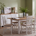 A white wooden dining table set with six matching slatted chairs with beige upholstered seats, styled in a dining room with a white sideboard, green walls, and potted ferns.