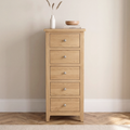 A tall, narrow five-drawer chest of light oak wood stands against a beige wall on a wood floor. The chest has a natural oak finish with small round silver knobs.