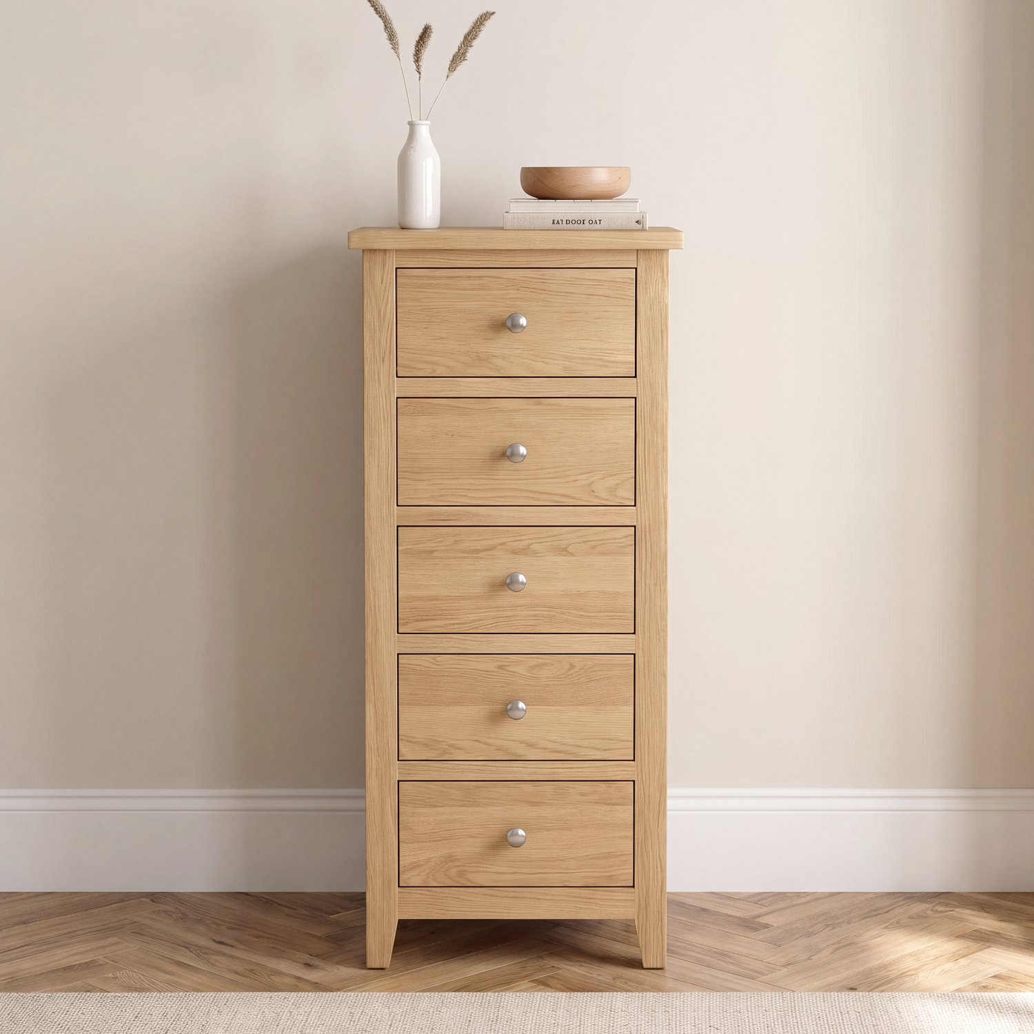 A tall, narrow five-drawer chest of light oak wood stands against a beige wall on a wood floor. The chest has a natural oak finish with small round silver knobs.
