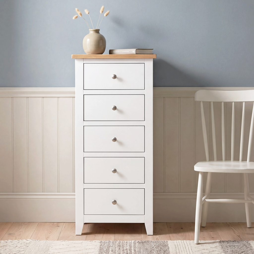 A white five-drawer chest of drawers with a light natural wood top stands against a blue-gray and white paneled wall, styled with a small vase and books, next to a white wooden chair.