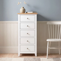 A white five-drawer chest of drawers with a light natural wood top stands against a blue-gray and white paneled wall, styled with a small vase and books, next to a white wooden chair.
