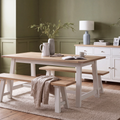 A light oak dining table with white painted trestle-style legs, accompanied by matching wooden benches, set in a styled dining room with green walls, a white sideboard, and a beige rug.