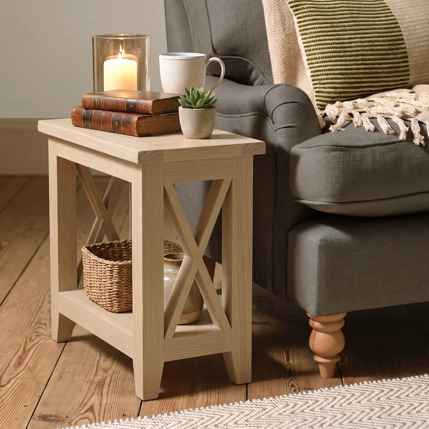 A light natural wood end table styled in a cozy living room next to a gray armchair, holding a lit candle, books, a white mug, and a small potted succulent.