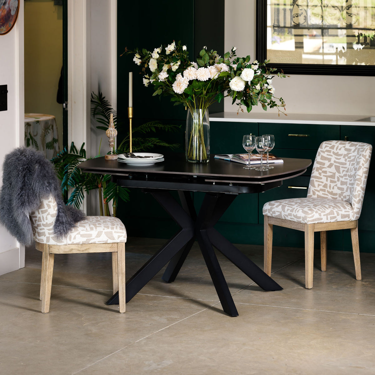 A modern dining table set in a dining room, featuring a black rectangular tabletop and a black X-shaped pedestal base, surrounded by two upholstered chairs with beige and white patterned fabric and light wood legs. The table is styled with a large vase of white flowers, wine glasses, and a candle.