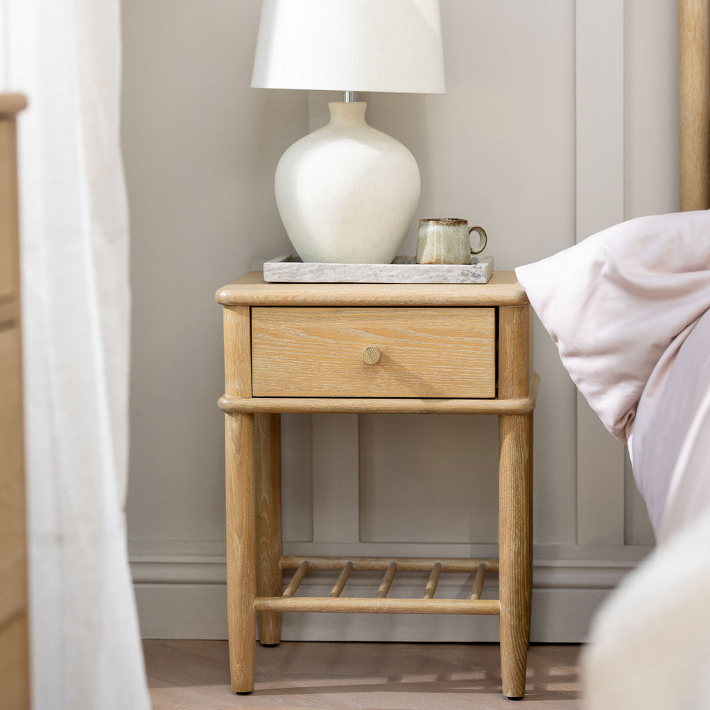 A light oak wood nightstand with a single drawer and open lower shelf, styled in a bedroom next to a bed with a white ceramic lamp and a small ceramic mug on top.