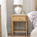 A light oak wood nightstand with a single drawer and open lower shelf, styled in a bedroom next to a bed with a white ceramic lamp and a small ceramic mug on top.