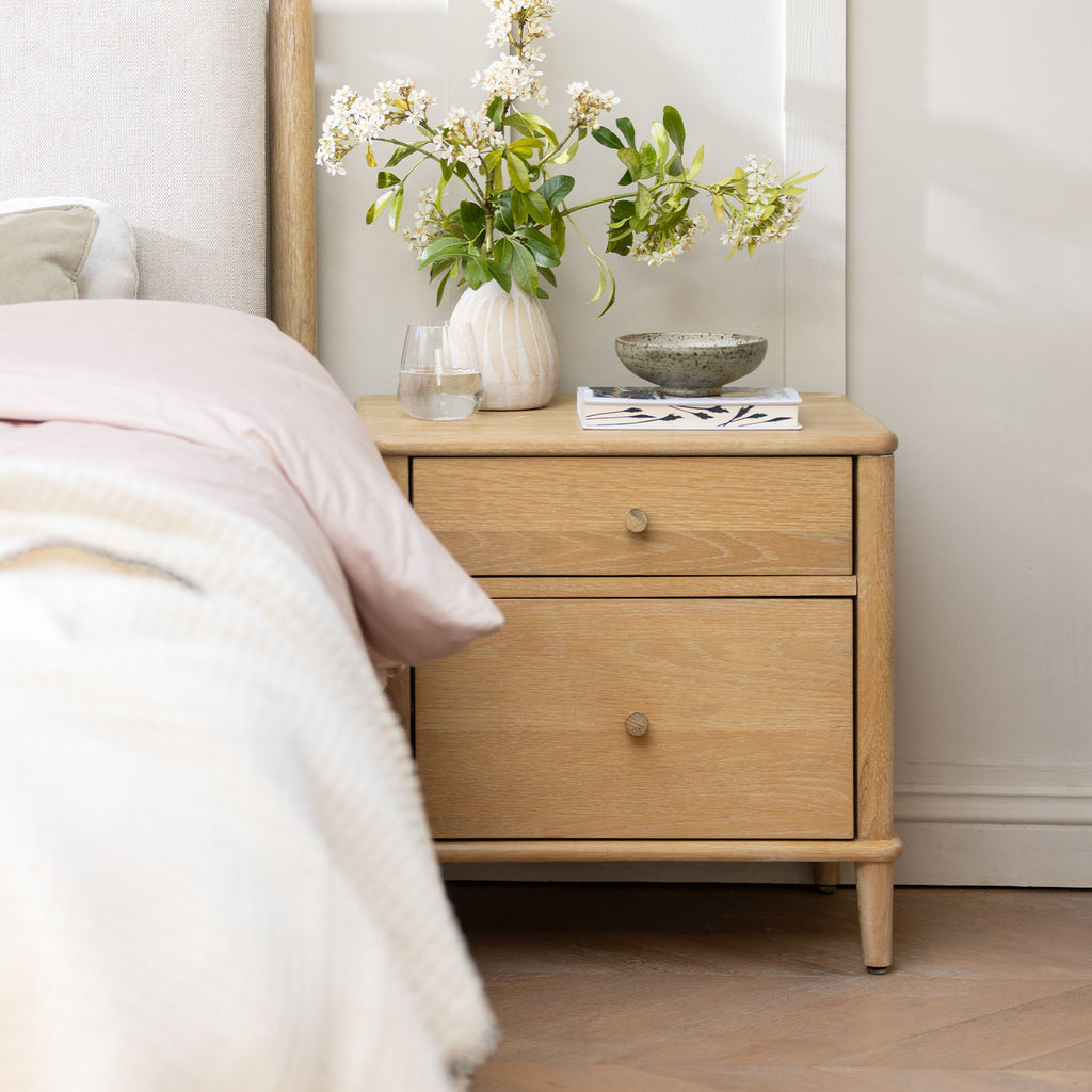 A light oak two-drawer nightstand in a bedroom setting, styled with a white ceramic vase of flowers, a small bowl, and a magazine next to a bed with pale pink bedding.