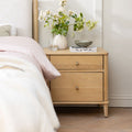 A light oak two-drawer nightstand in a bedroom setting, styled with a white ceramic vase of flowers, a small bowl, and a magazine next to a bed with pale pink bedding.