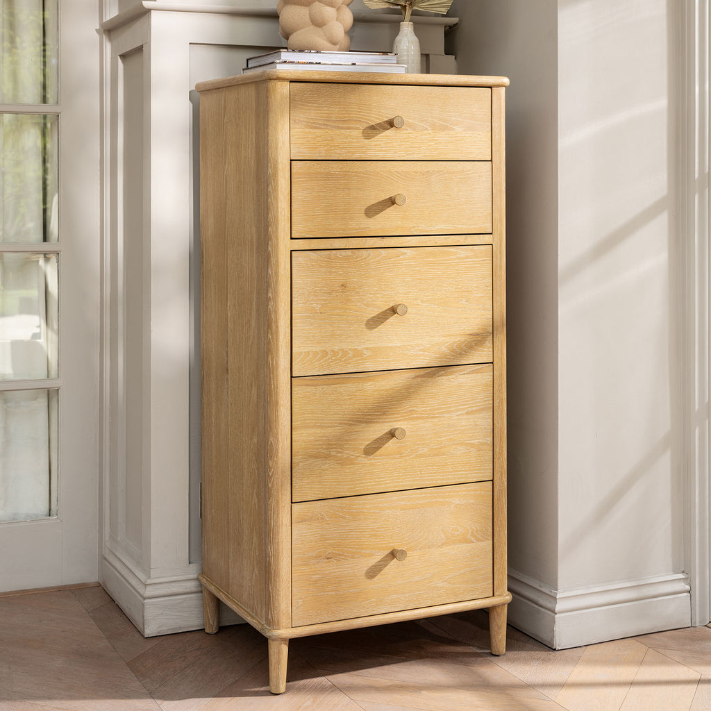 A tall, narrow wooden chest of drawers with five drawers and small round knobs, shown in a sunlit room corner against white walls.