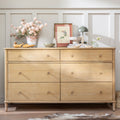 A light natural wood six-drawer dresser with round knobs, styled with home decor items including a vase of pink flowers, a green lamp, and books, set against a white paneled wall.