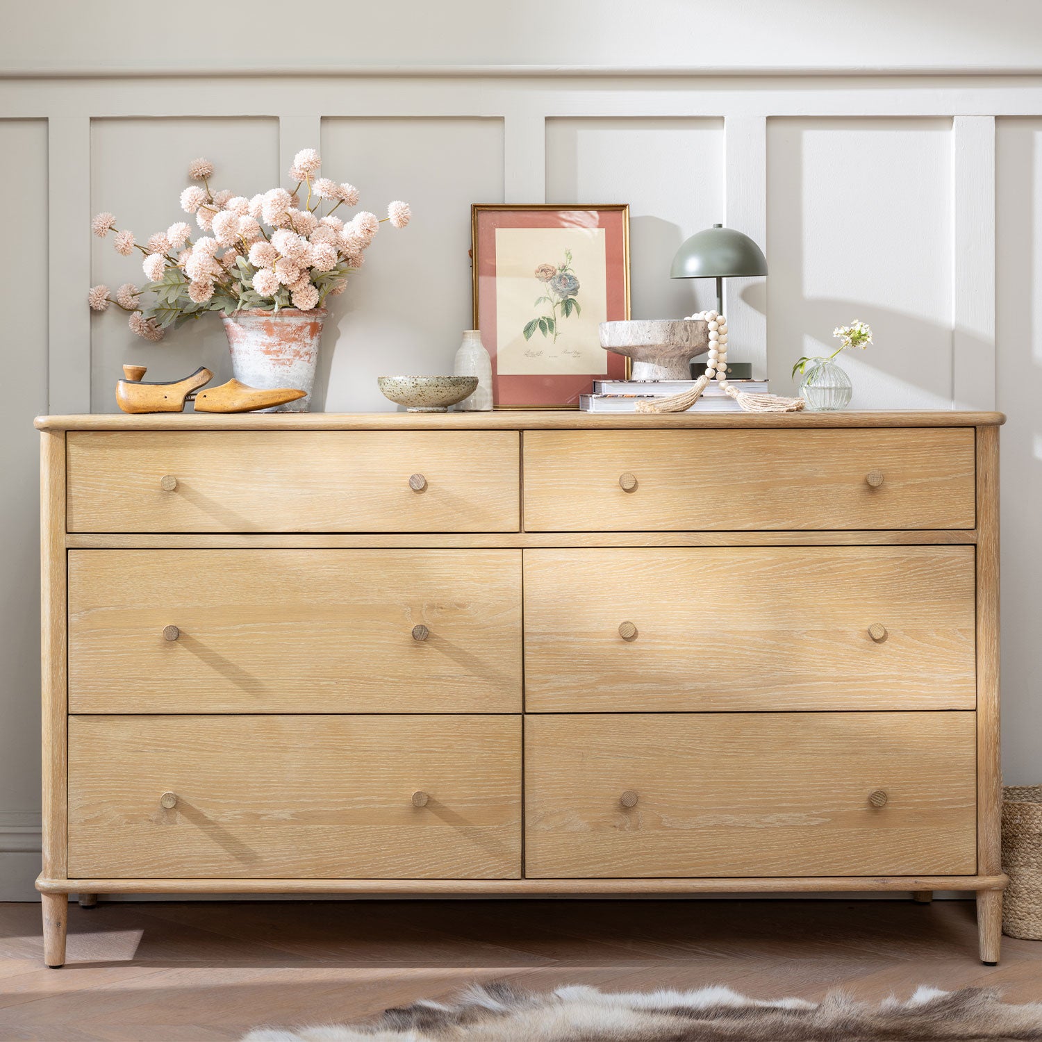 A light natural wood six-drawer dresser with round knobs, styled with home decor items including a vase of pink flowers, a green lamp, and books, set against a white paneled wall.