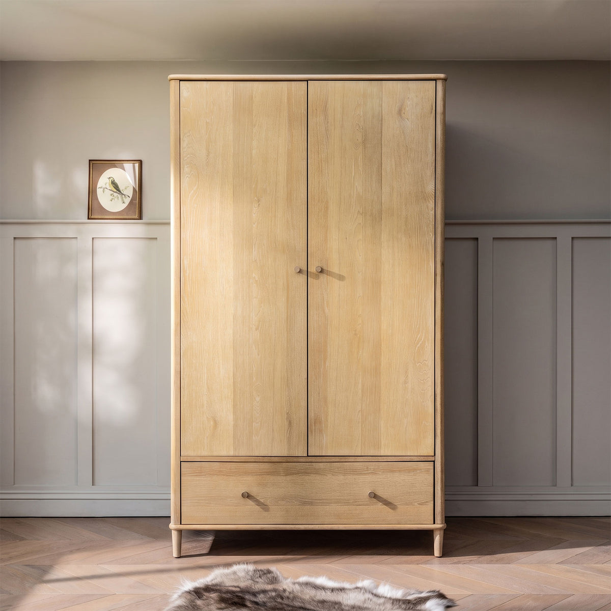 A tall light oak double-door wardrobe with a bottom drawer, set against a grey paneled wall and wooden floor.