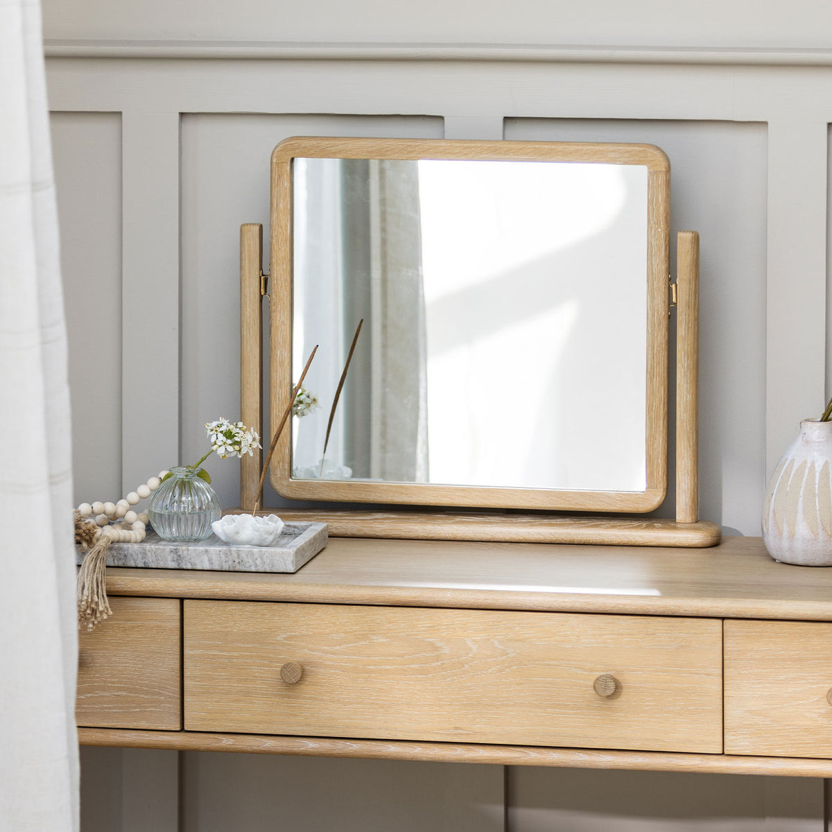 A light oak wood folding vanity mirror with a rectangular center mirror and two hinged side mirrors, displayed on a matching wooden dresser against a gray paneled wall.