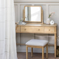 A light natural wood vanity table with three drawers, a matching folding mirror, and a cushioned stool, styled in a bright room with a white curtain, vases, and pink flowers.