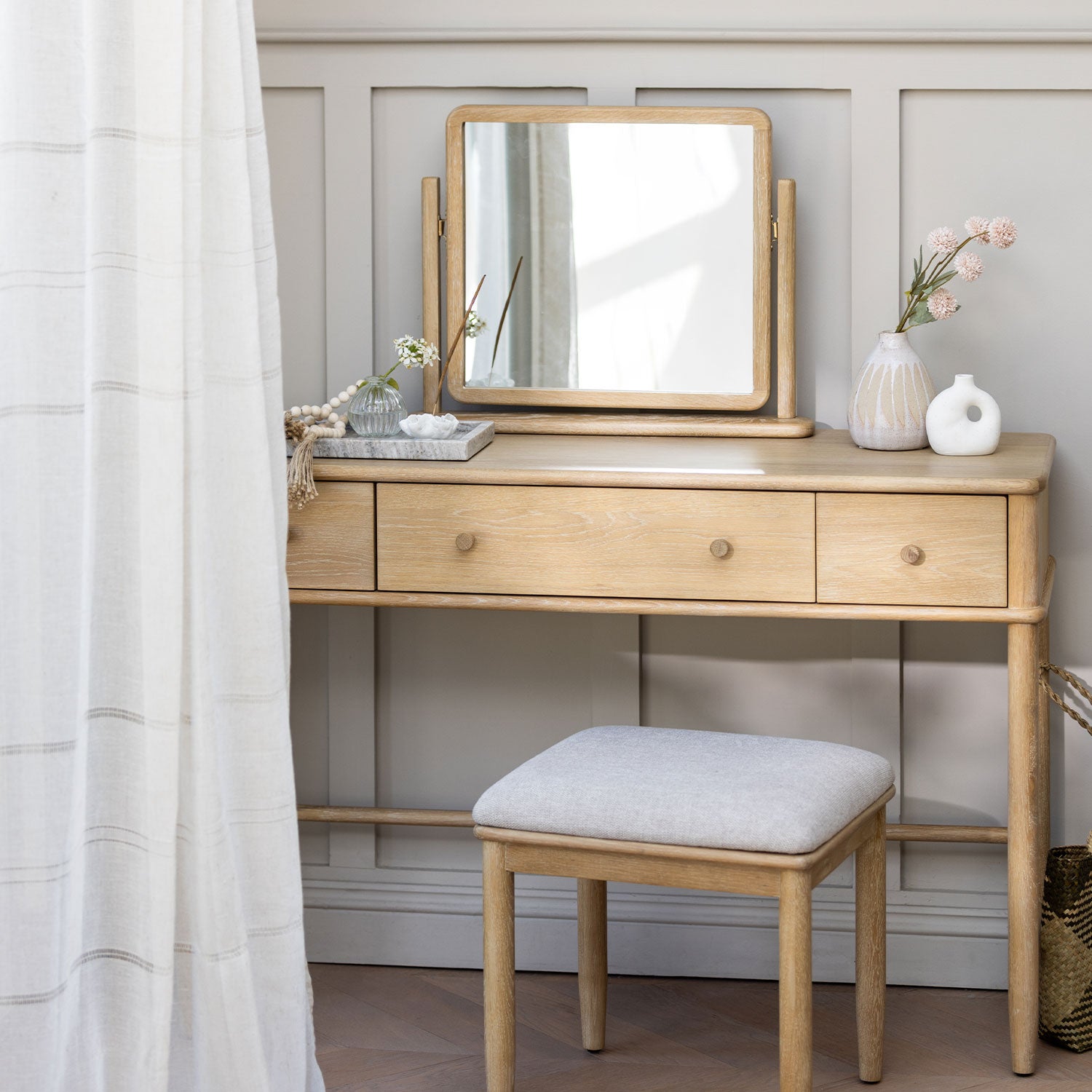 A light natural wood vanity table with three drawers, a matching folding mirror, and a cushioned stool, styled in a bright room with a white curtain, vases, and pink flowers.